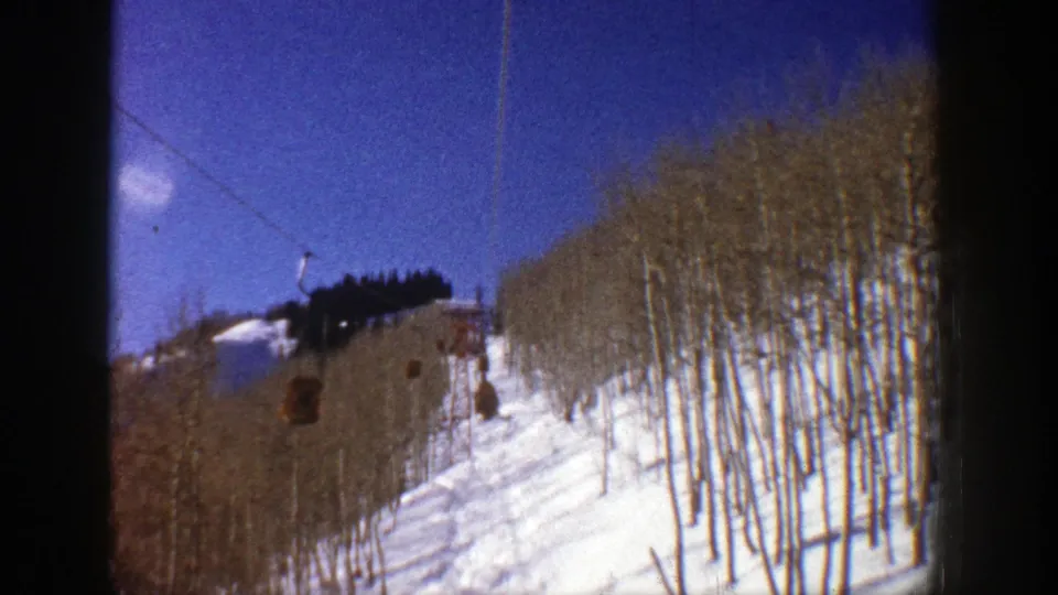 Visitor-shot 8mm view in Aspen showing ski lift cabins approaching over a snow-covered hillside and bare trees.