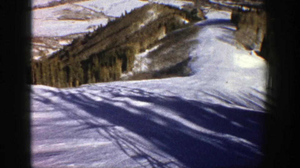 1961 Aspen winter frame showing a snow-covered slope or trail with long tree shadows and surrounding mountain terrain.