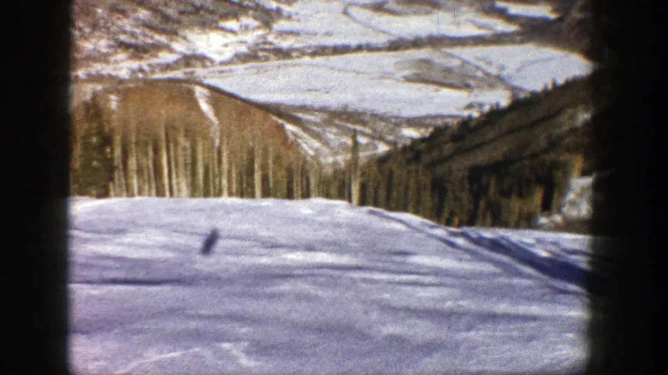 1961 Aspen ski-slope frame with a small distant skier crossing the snow below wooded hills and mountain ridges.