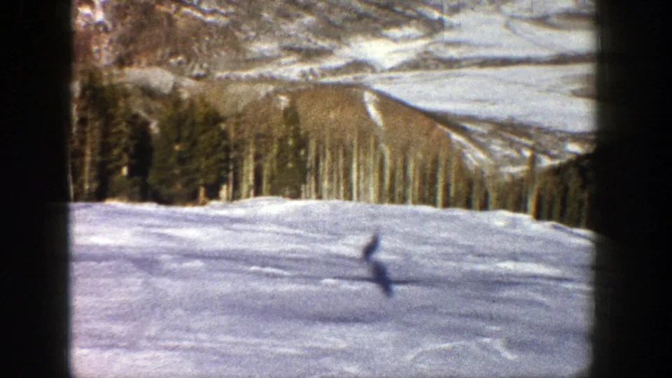 Vintage Aspen frame showing a lone distant skier on a broad snowy run framed by trees and winter mountain landscape.