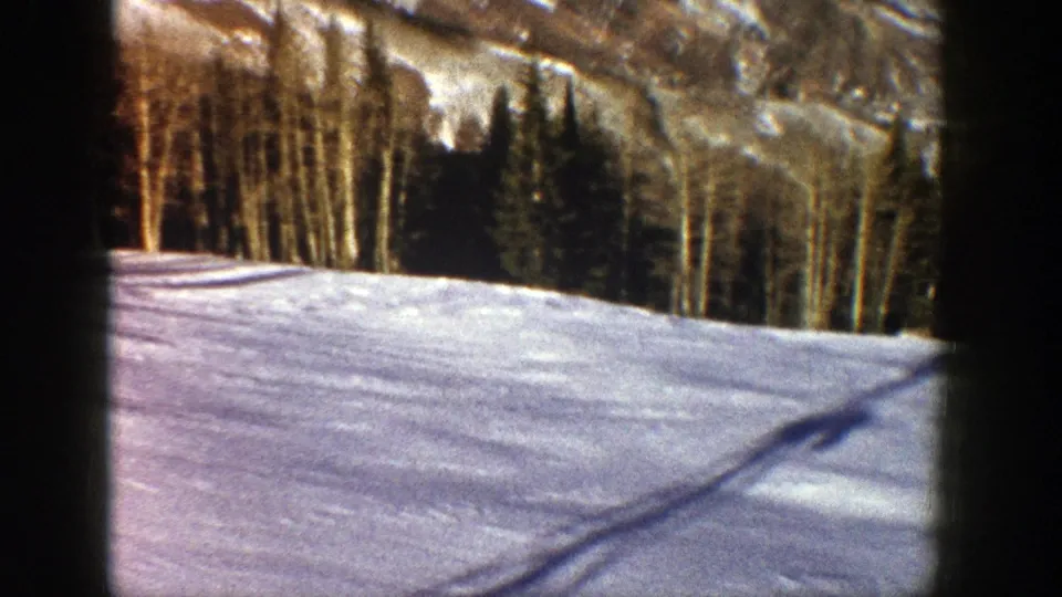 1961 Aspen, Colorado frame of a quiet snow-covered field, elongated shadows, tree line, and rugged mountain slope beyond.