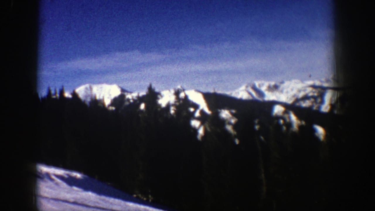 1961 Aspen-area 8mm view of snow-covered mountain peaks rising behind a dark evergreen forest under a blue sky.