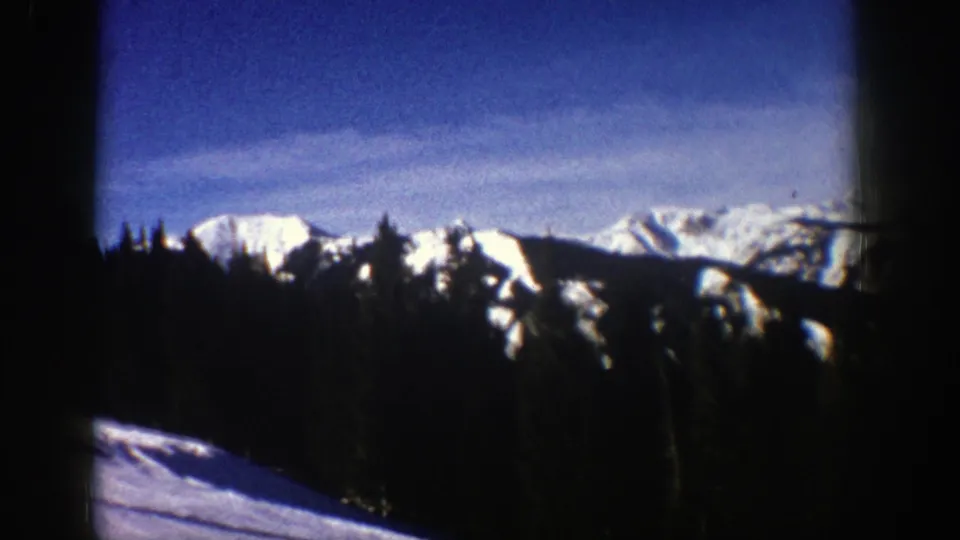 1961 Aspen-area 8mm view of snow-covered mountain peaks rising behind a dark evergreen forest under a blue sky.