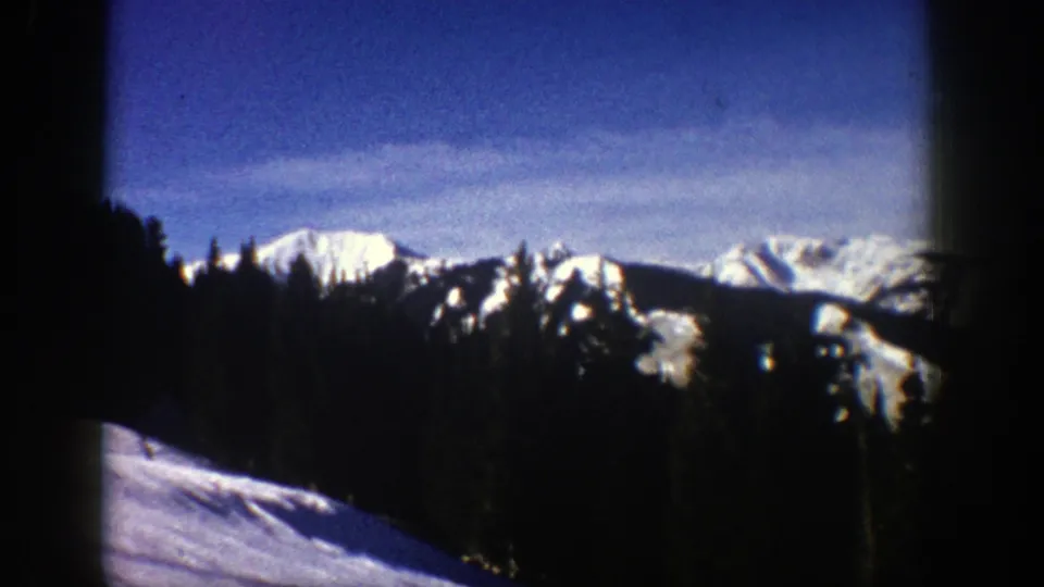 Vintage 8mm frame from Aspen, Colorado, showing snow-bright mountain ridges beyond dark trees and a sloping snowy foreground.