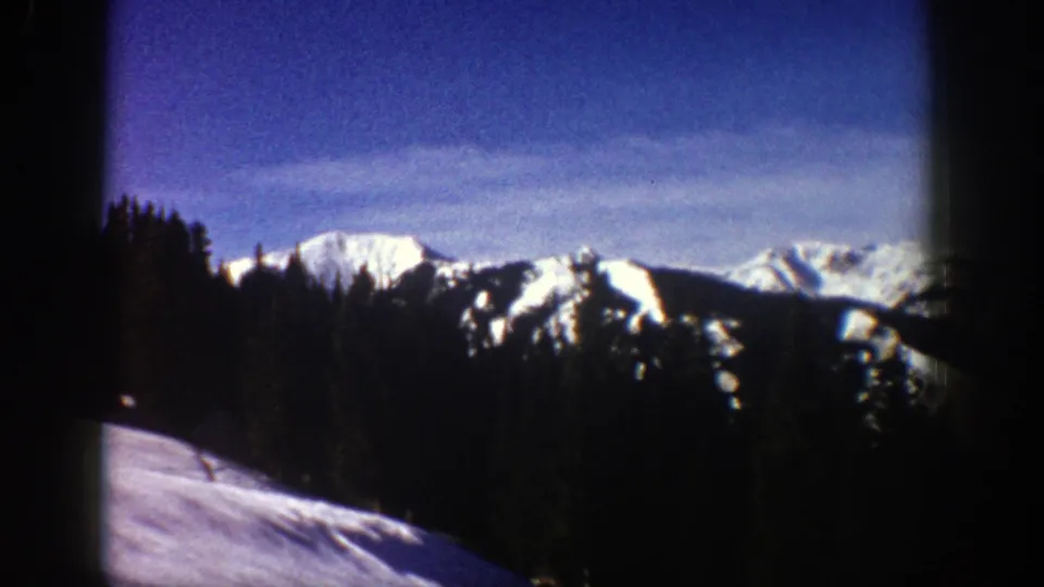 1961 Aspen-area 8mm frame showing snowy peaks, dense evergreen forest, blue sky, and a dark branch near the right edge.