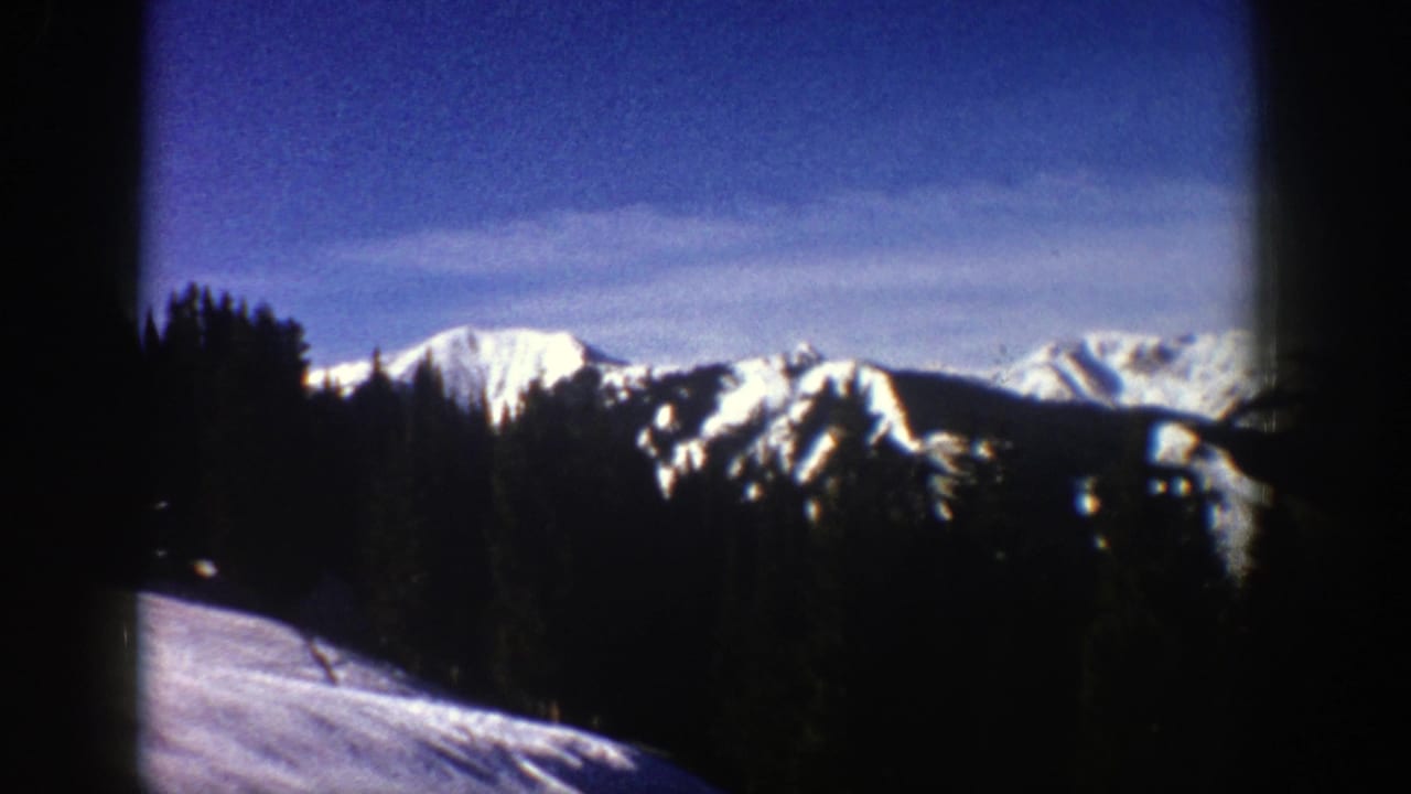 1961 Aspen-area home-movie frame showing snow-covered mountain ridges partly framed by dark evergreens and film-edge shadow.