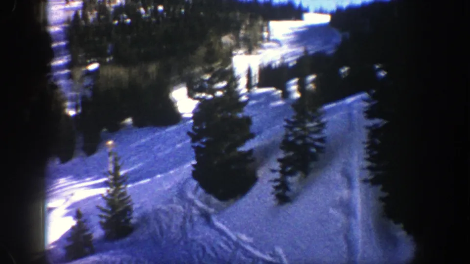 1961 Aspen home-movie frame showing a snow-covered slope, scattered evergreens, curved tracks, and dark film-edge shadow.