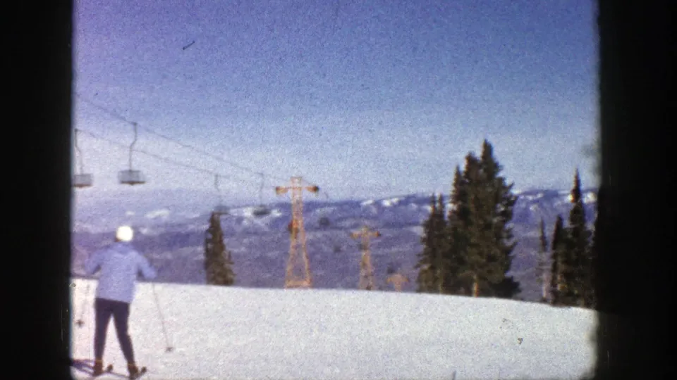 1961 Aspen frame showing a skier at the edge of a snowy slope, with lift towers and distant mountain ridges beyond.