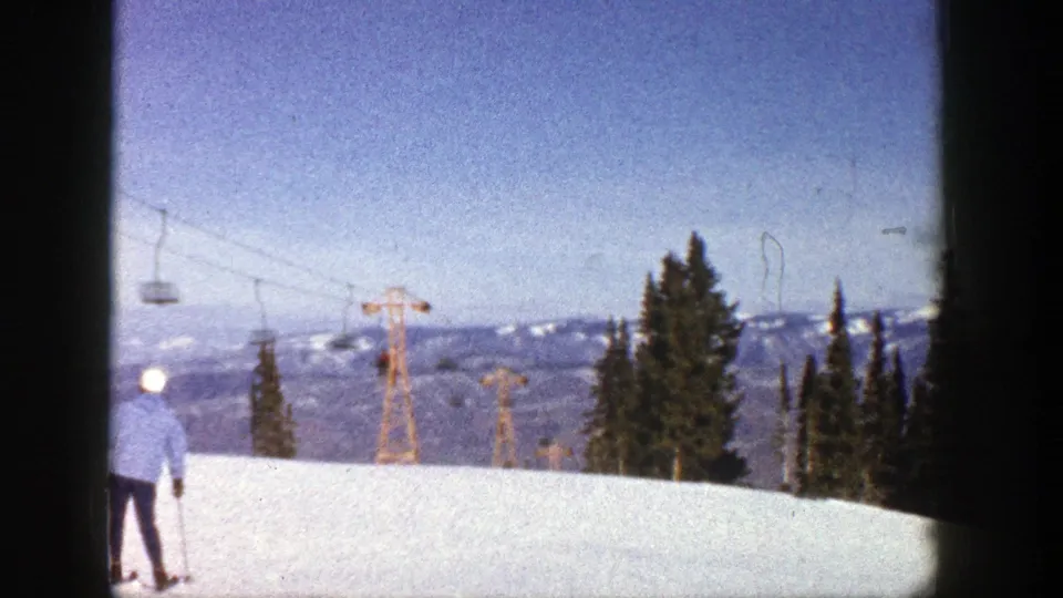 1961 Aspen ski-area view with a distant skier, chairlift structures, evergreen trees, and blue mountain horizon.