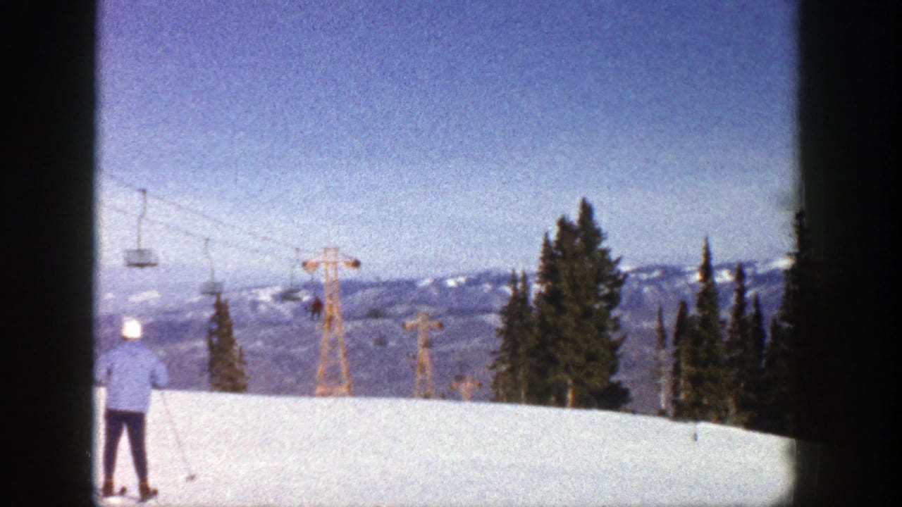 1961 Aspen ski-slope view with a skier in a blue jacket near the frame edge, chairlift towers, evergreens, and distant mountains.