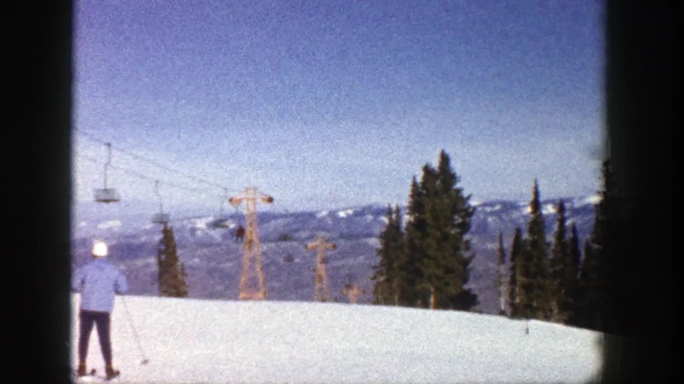 1961 Aspen ski-slope view with a skier in a blue jacket near the frame edge, chairlift towers, evergreens, and distant mountains.