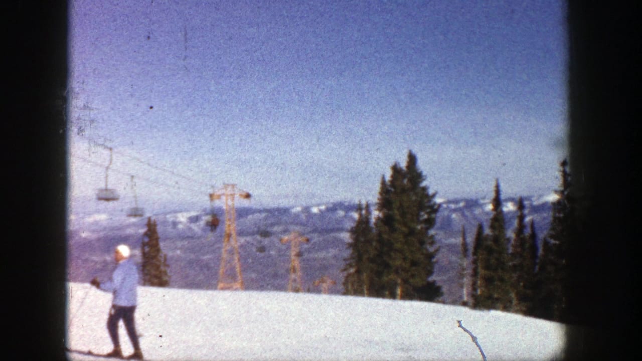 Skier in blue stands on snow facing chairlifts, with evergreens and mountains in the distance.