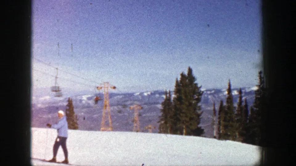 A 1961 Aspen home-movie frame shows the skier lower on the slope, with chairlift machinery, evergreens, and distant snow-covered mountains.