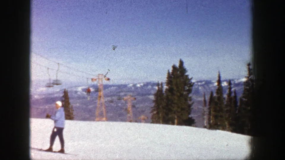 The skier remains on the snow in this Aspen 1961 frame, set against chairlift towers, conifers, and a pale mountain horizon.