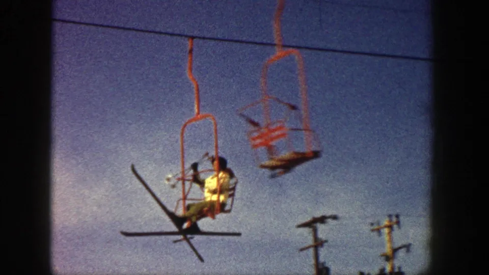 Aspen, Colorado 1961 home-movie frame showing a skier on a red chairlift with skis angled outward against blue sky.