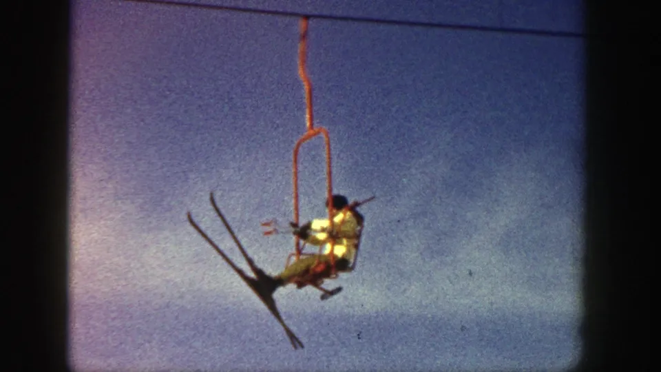Home-movie frame from Aspen in 1961 showing a skier in a light jacket riding a red chairlift above the camera.