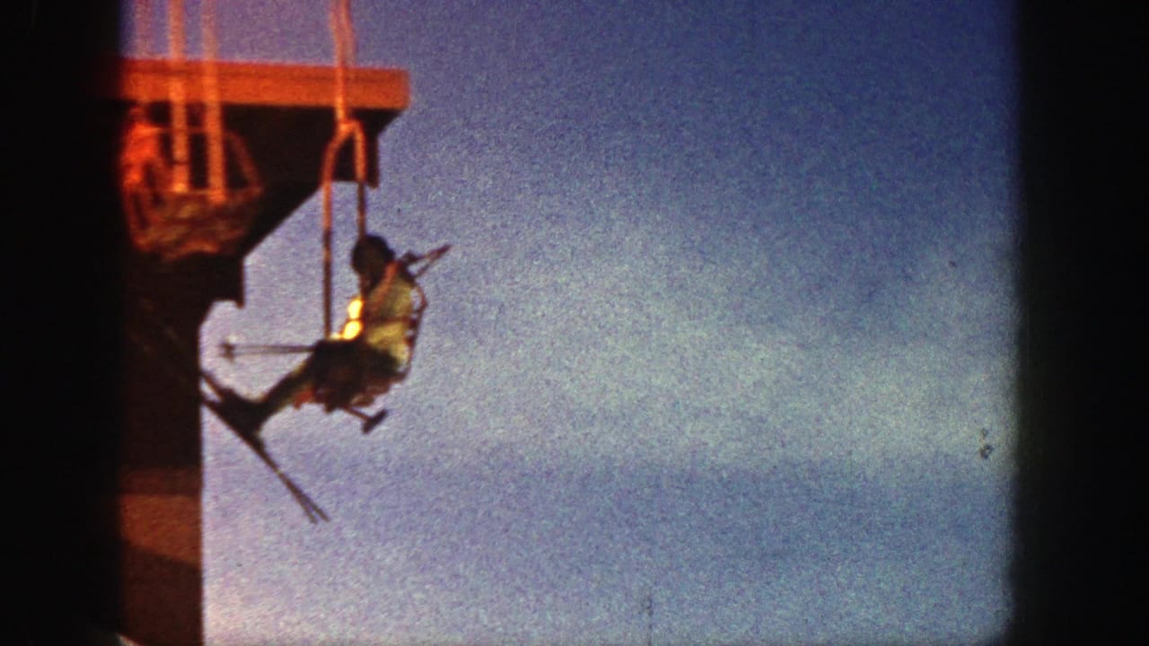 1961 Aspen home-movie frame showing a skier on a chairlift with skis raised near a dark lift structure and blue sky.