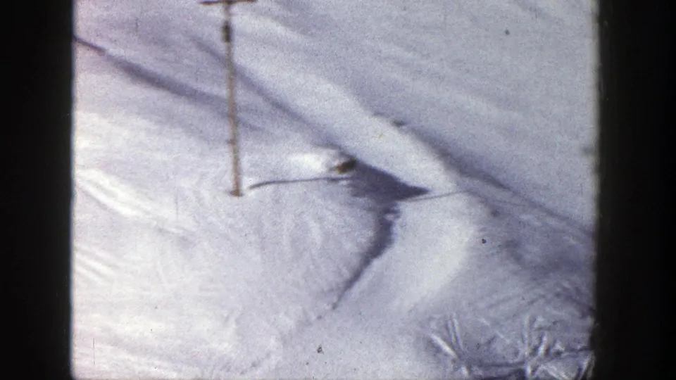 The frame looks down at a snow-covered ski slope with a pole, tracks, and wind-shaped surface texture.
