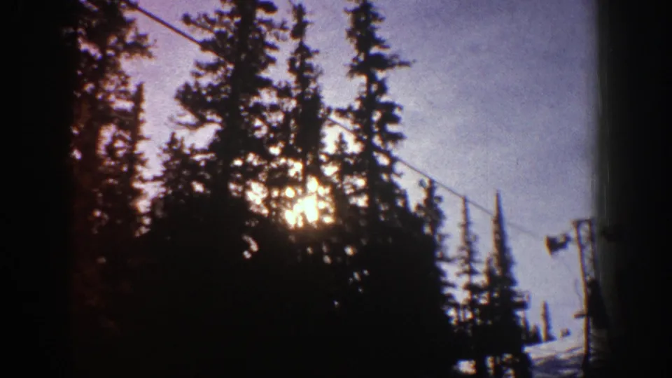 Aspen ski-resort home-movie frame showing low sun behind evergreens and a lift cable crossing the winter scene.