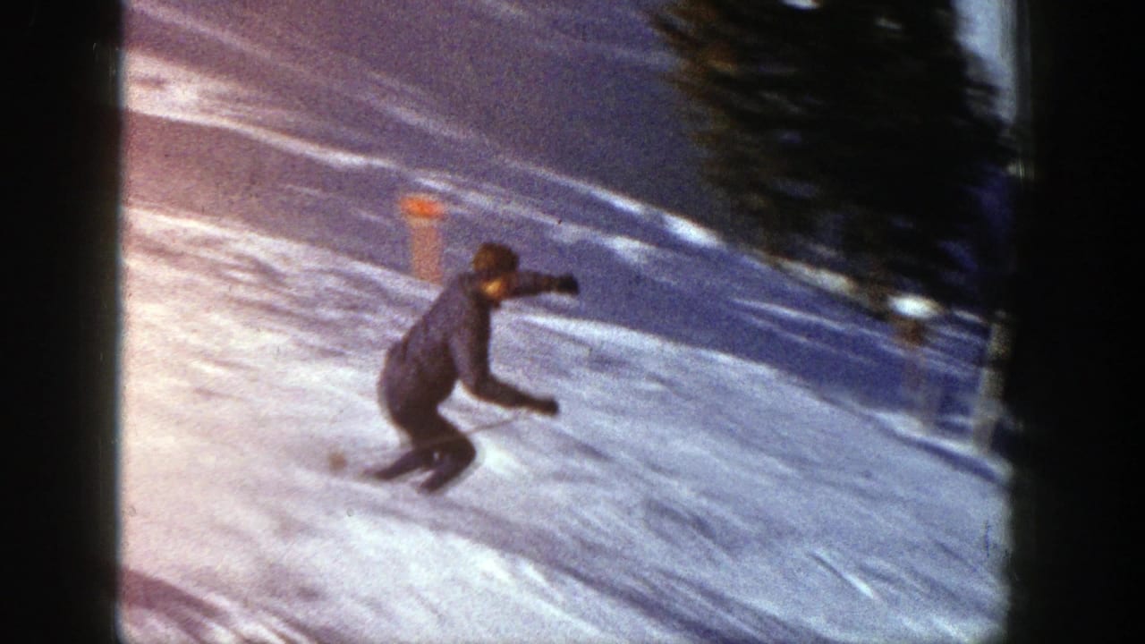 1961 Aspen 8mm frame of a skier in a dark jacket carving across a bright snowy slope near trees.