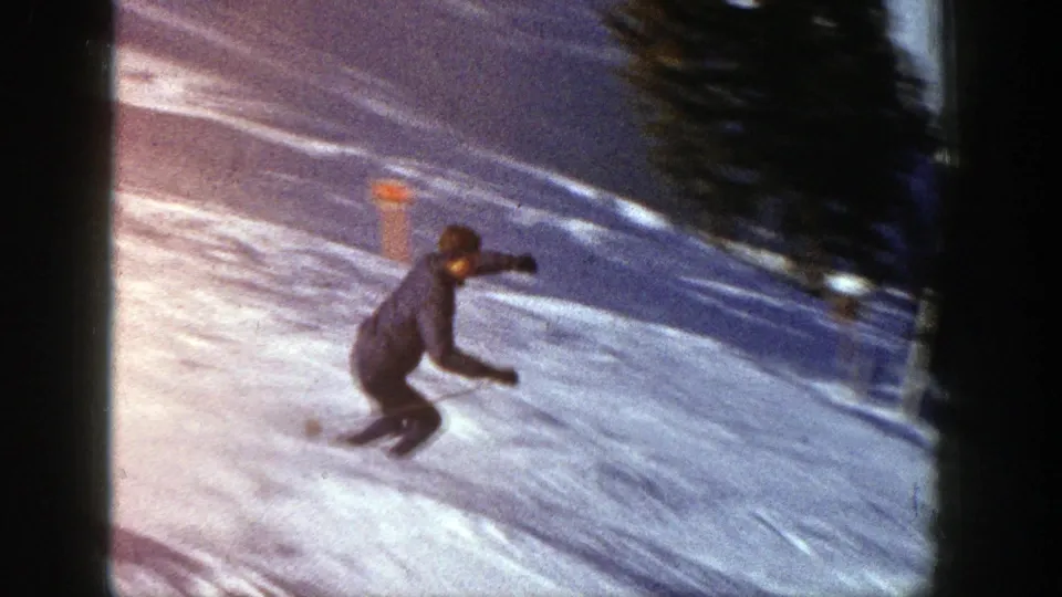 1961 Aspen 8mm frame of a skier in a dark jacket carving across a bright snowy slope near trees.