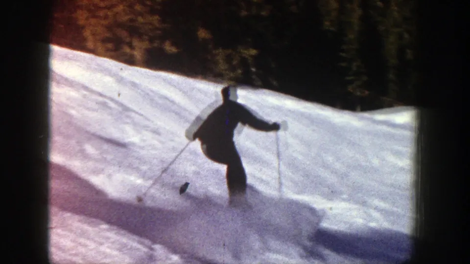 Backlit 8mm view of a skier moving away on an Aspen slope, with bright snow and dark evergreens behind.