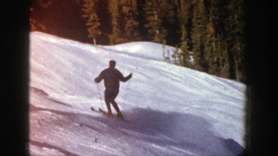 1961 Aspen home-movie frame showing a skier from behind on a wide snowy slope bordered by evergreens.