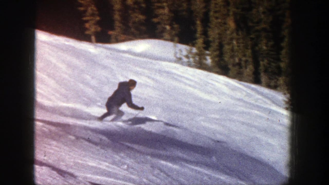 A skier turns downhill on a snowy Aspen, Colorado slope in 1961, seen in soft 8mm home-movie color with trees behind.