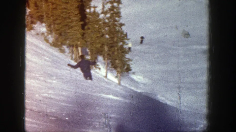 A distant skier descends through the lower part of the frame, with snowy ground and tall evergreens visible in the 8mm image.