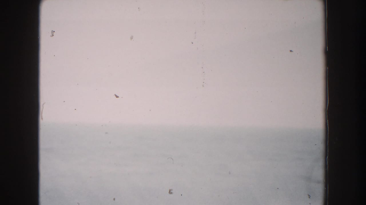 1960 Italian home-movie frame showing a hazy sky and faint sea horizon, softened by 8mm grain and exposure variation.