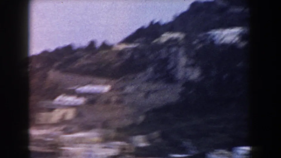 1960 Italian home-movie frame of a rugged hillside with scattered light buildings, softened by handheld blur and 8mm grain.