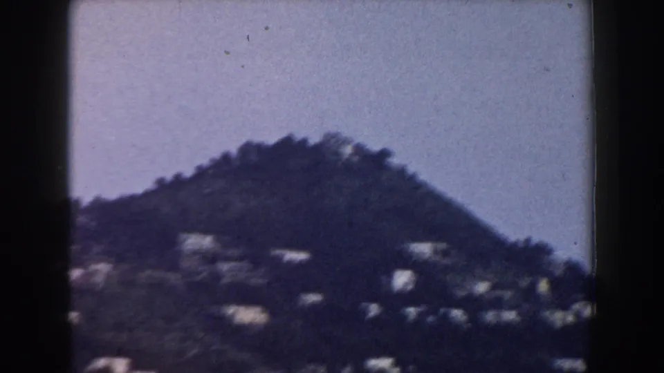 Distant Italian mountain slope in a 1960 home-movie frame, with scattered buildings below a grainy, pale sky.