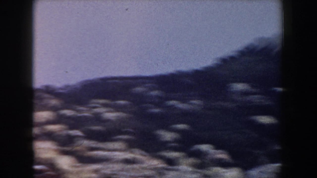 1960 Italy home-movie frame showing a soft, distant hillside settlement near the sea under a hazy purple-blue sky.