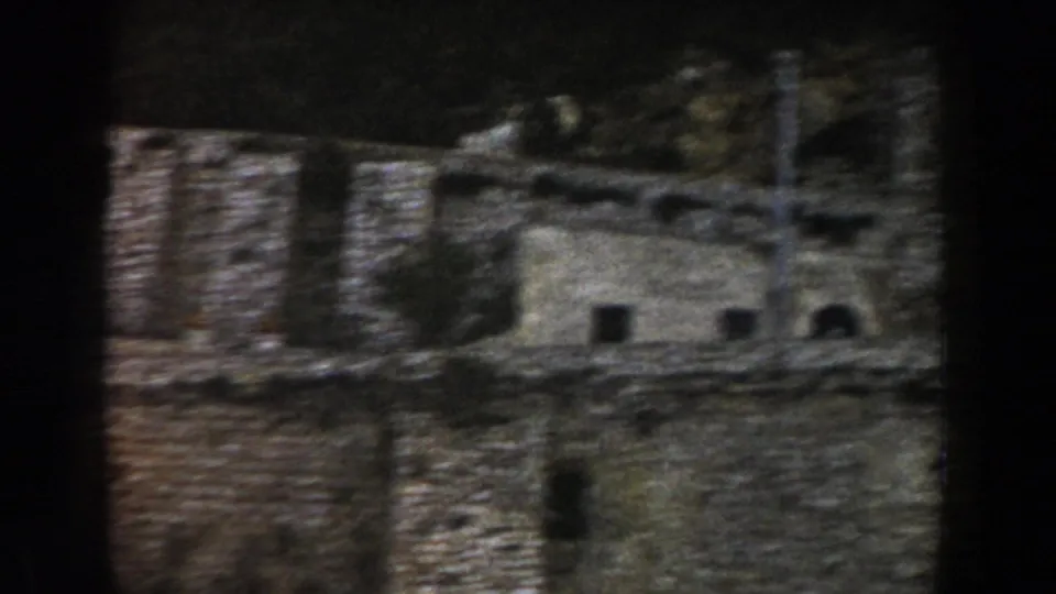 1960 Italy frame showing rough stone walls and building openings against a dark wooded slope.