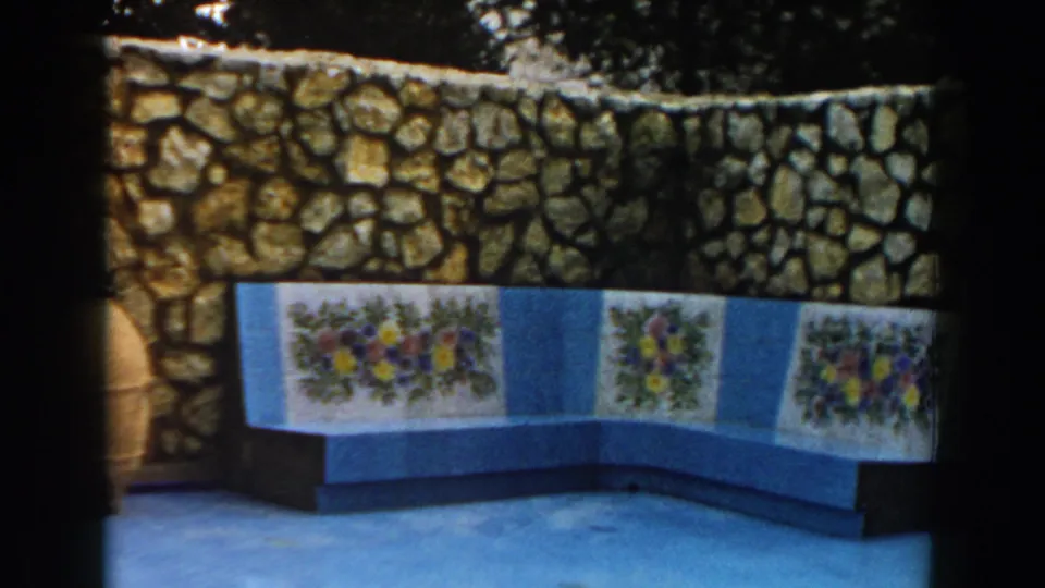 1960 Italy travel frame showing a stone wall and blue built-in bench decorated with floral tile panels outdoors.