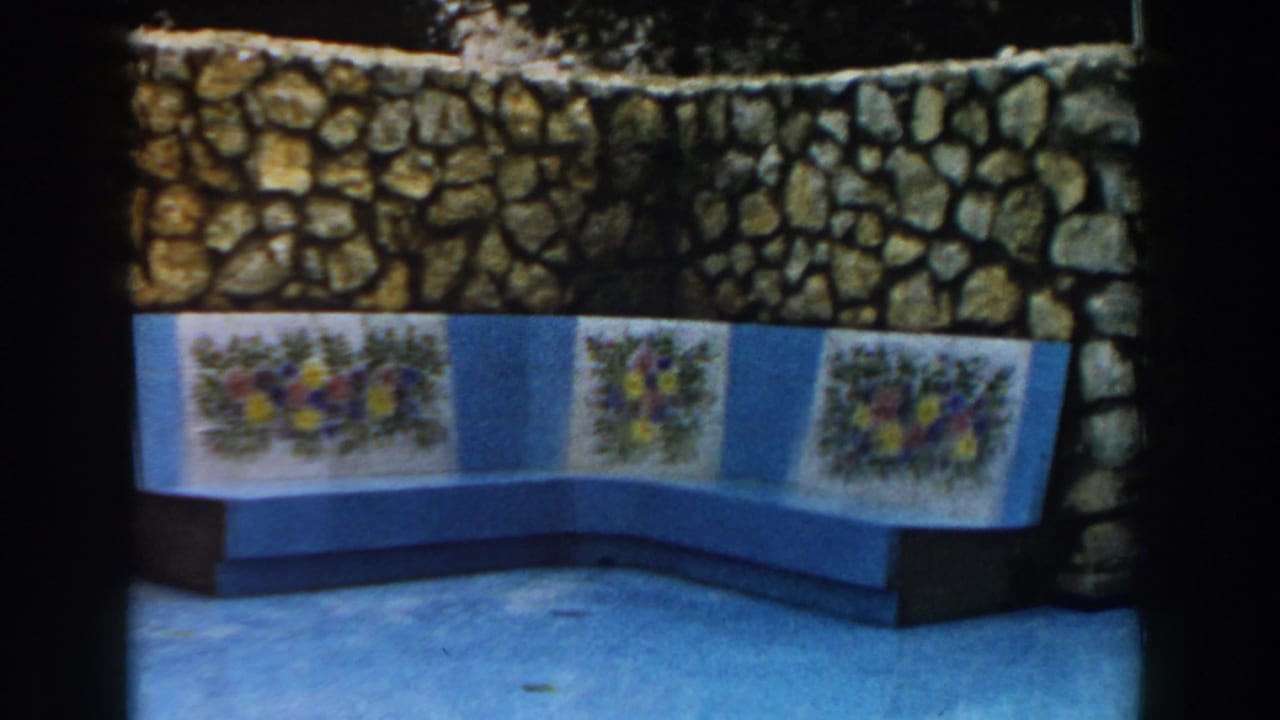 1960 Italy 8mm frame showing a blue built-in bench, floral tile panels, tan stone wall, and reflective pool paving.