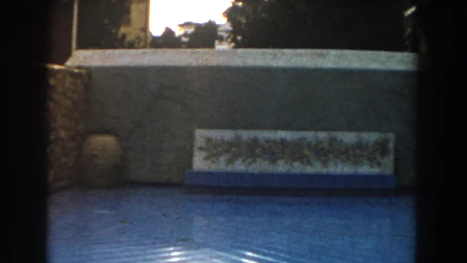 1960 Italy 8mm frame showing a low wall, floral tile panel, stone edge, and bright reflections across a blue pool surface.