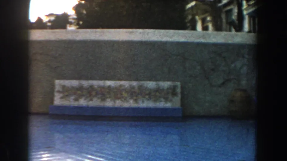 1960 Italy home-movie frame with a floral tile panel, blue reflective pool surface, stonework, and a small urn near the wall.