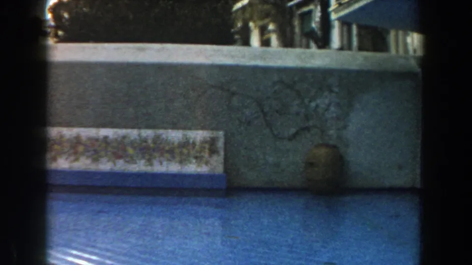 1960 Italy home-movie frame holding on a courtyard wall with a tile panel, blue foreground surface, and earthenware jar.