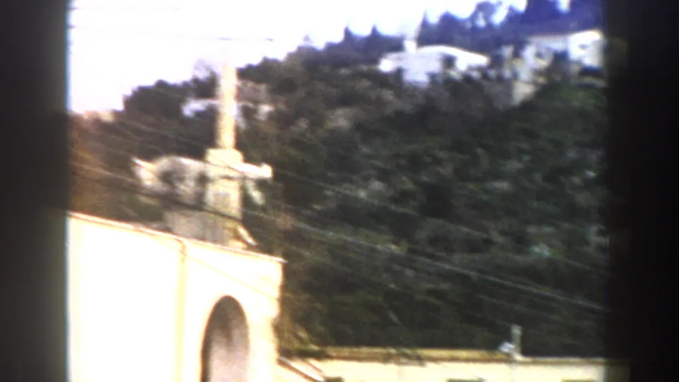 1960 Italy 8mm frame showing a tree-covered hillside with pale houses, utility lines, and a white arched structure in the foreground.