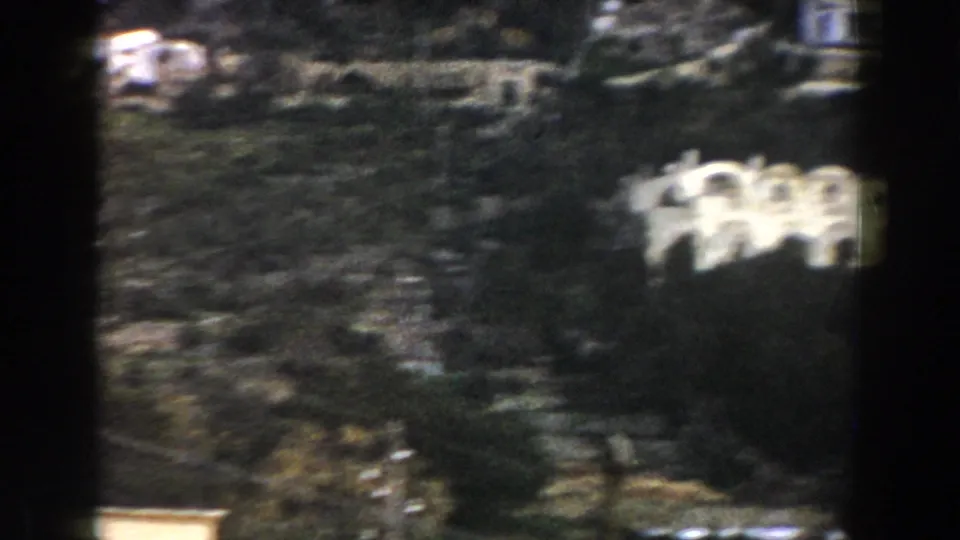 1960 Italy home-movie frame of a wooded hillside, scattered pale buildings, and a white arched structure partly obscured by soft focus.