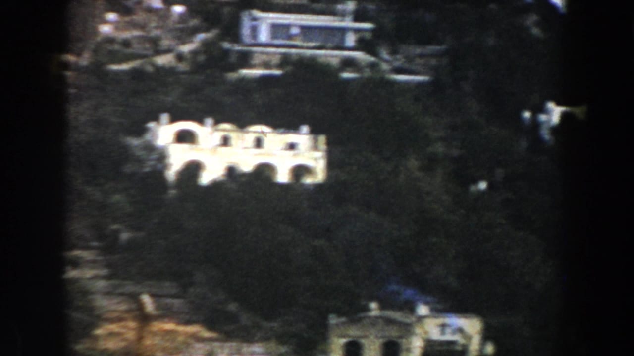 White arched building on a wooded hillside with other structures above.