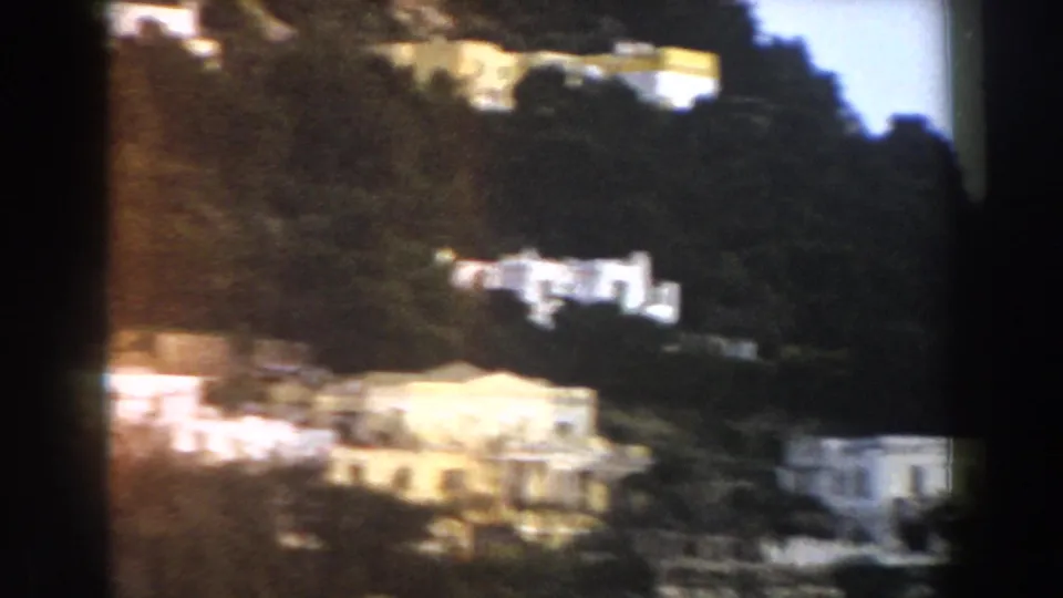 Soft 8mm view of pale buildings set across an Italian hillside, with trees and terraces partly obscuring the structures.