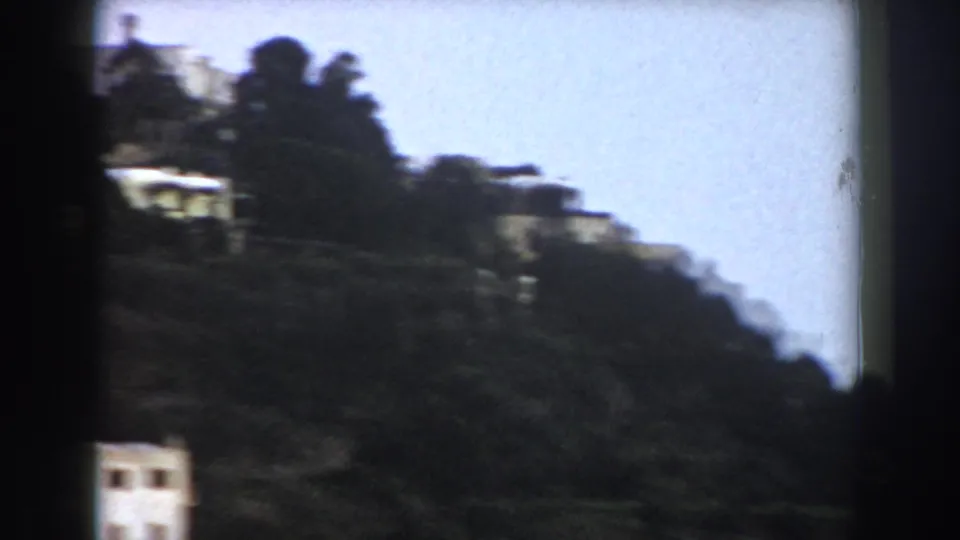 The pan reaches a darker wooded slope with a small white structure, terracing, and pale sky along the hillside edge.