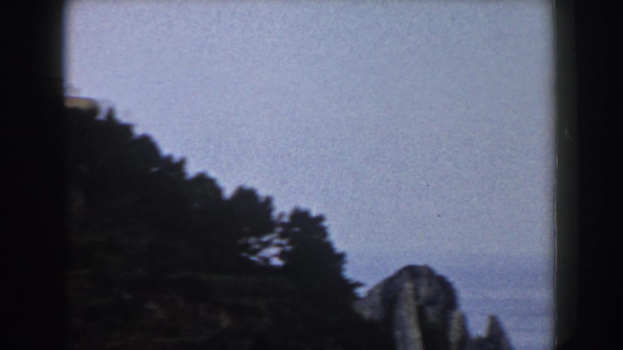 1960 Italy 8mm frame with dark trees, a pale sky, and a distant coastal cliff or rock form.