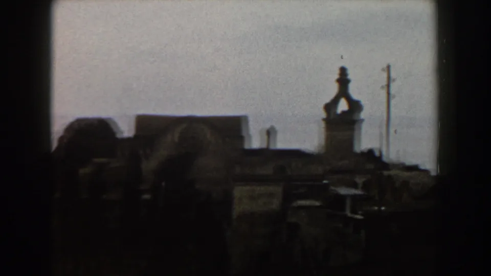 1960 Italy home-movie frame showing shadowed rooftops, a tower-like structure, utility pole, pale sky, and distant sea.