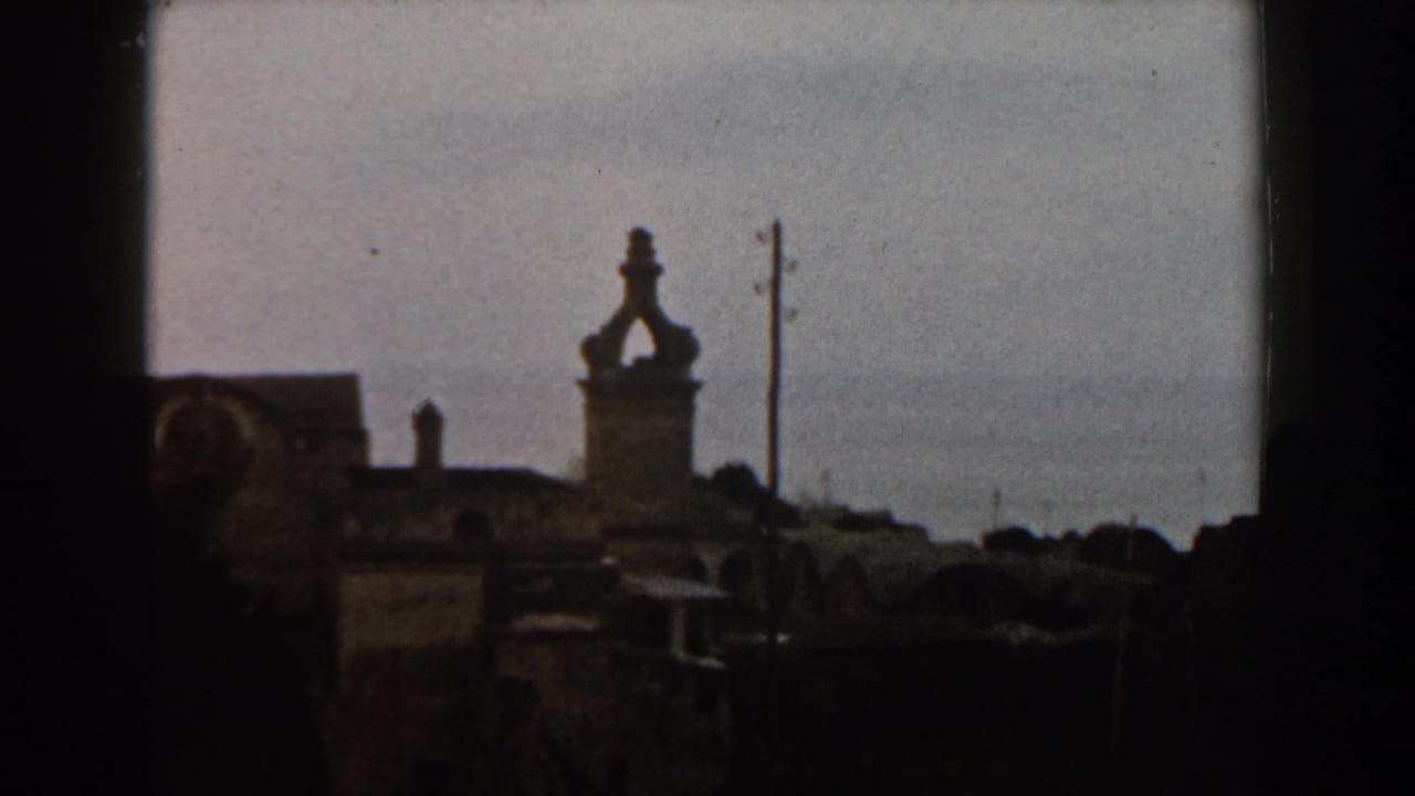 Town rooftops with a tower, utility pole, pale sky, and sea horizon.