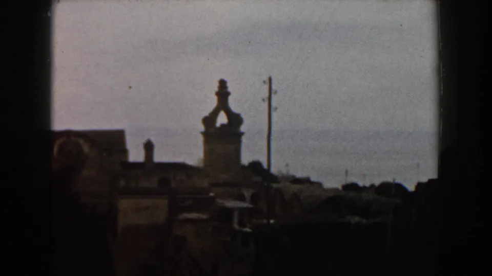 1960 Italy frame of weathered town buildings, a tower form, utility pole, and soft sea horizon in the background.