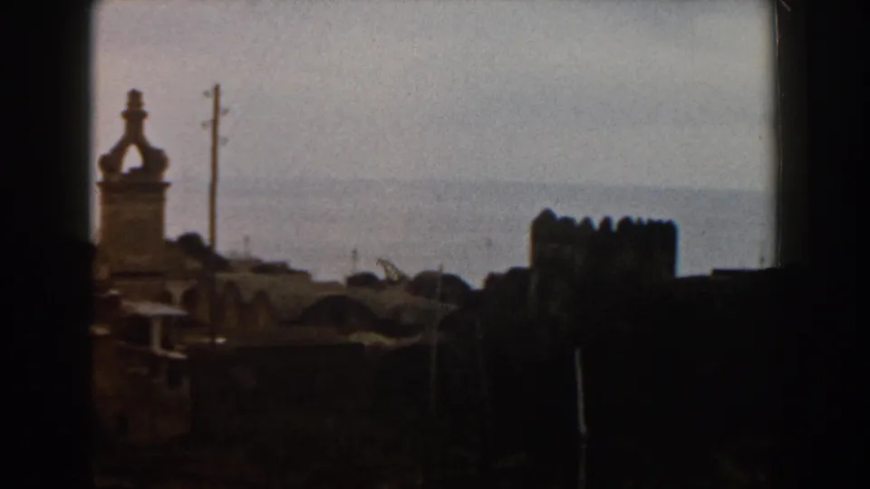 1960 Italy home-movie still showing a tower at left, a utility pole, dark rooftops, and a crenellated structure near the sea.