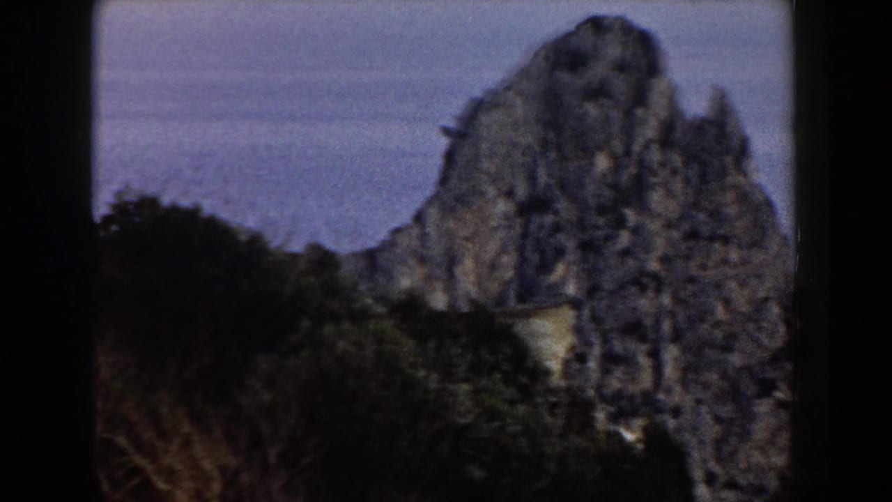 1960 Italy 8mm frame showing a tall weathered rock mass beside the sea, with dark vegetation at the base and soft blue-gray water behind it.